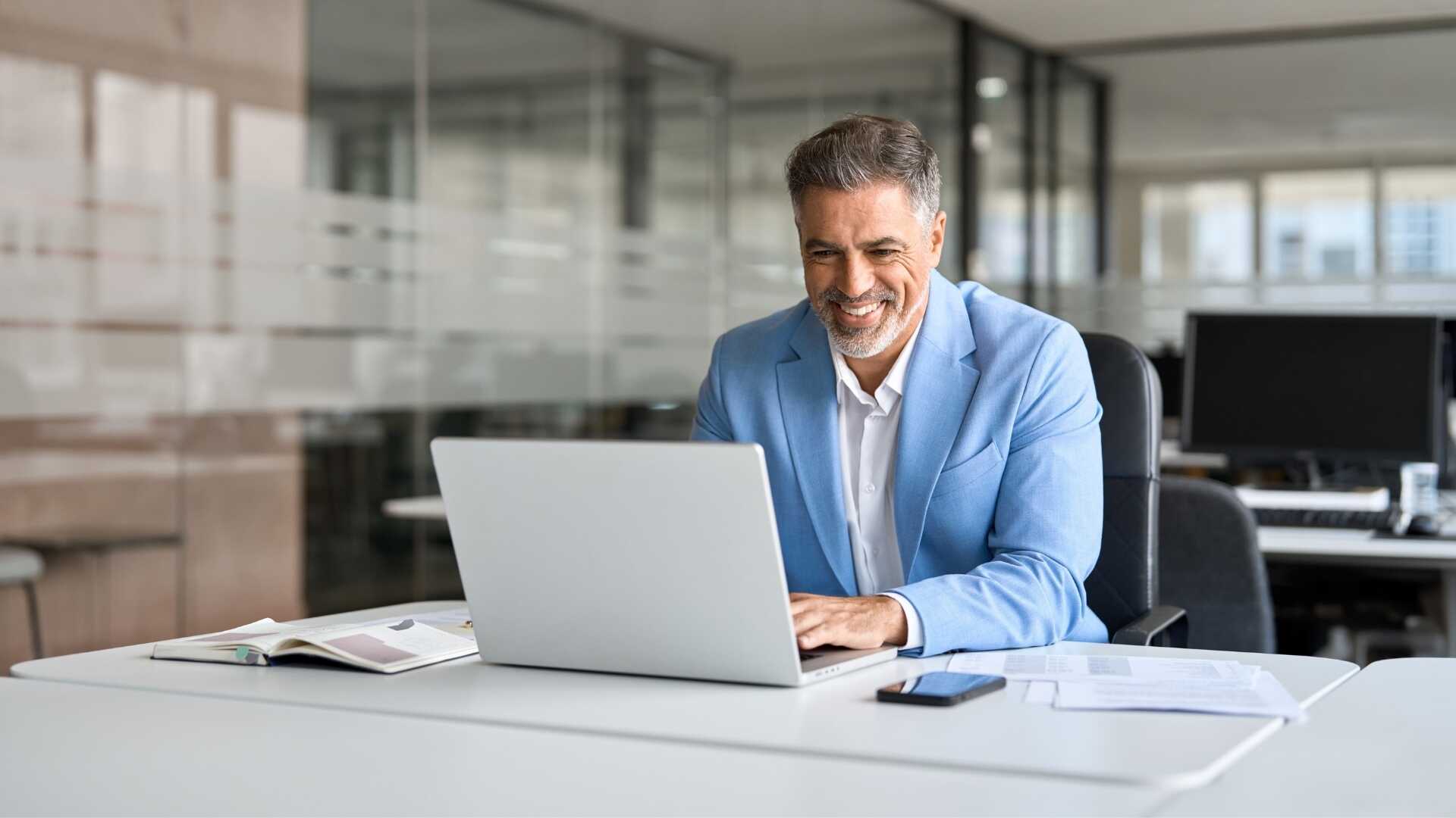 Confident business professional in a white shirt and light blue blazer working on his laptop in a modern office.